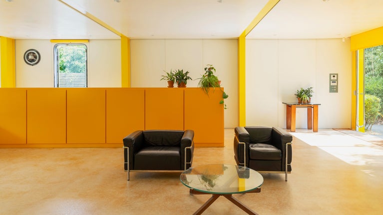 Two black armchairs and a round glass coffee table sit in front of a bright yellow cabinet. Sunlight shines thought the full height windows.